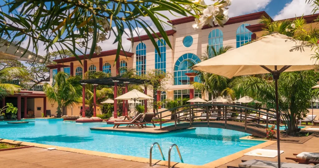 Tropical hotel resort with a swimming pool, lounge chairs, and a footbridge in front of the main hotel building.